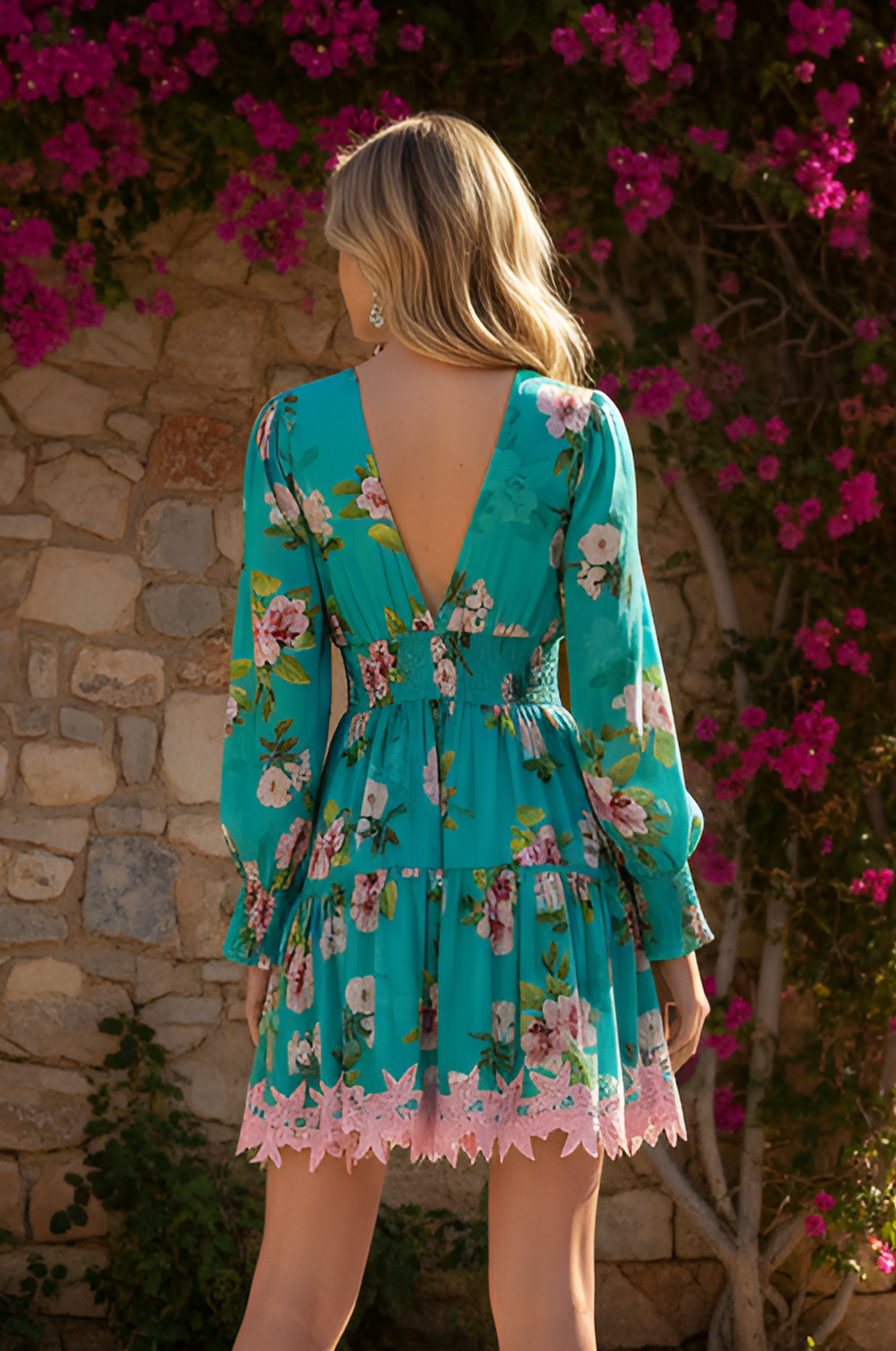 A blonde woman stands outdoors by a stone wall, surrounded by blooming pink flowers, wearing the Chloe Turquoise Floral Long-Sleeve Casual Dress.